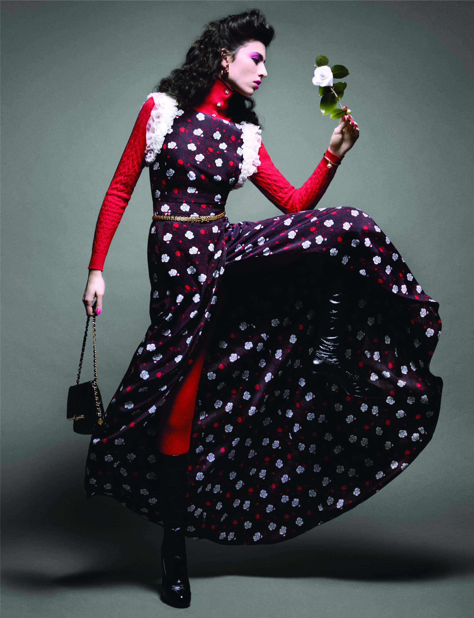 Woman in polka-dot dress holding a white rose in a studio shot. She also has black boots, red sweater, and a black purse.