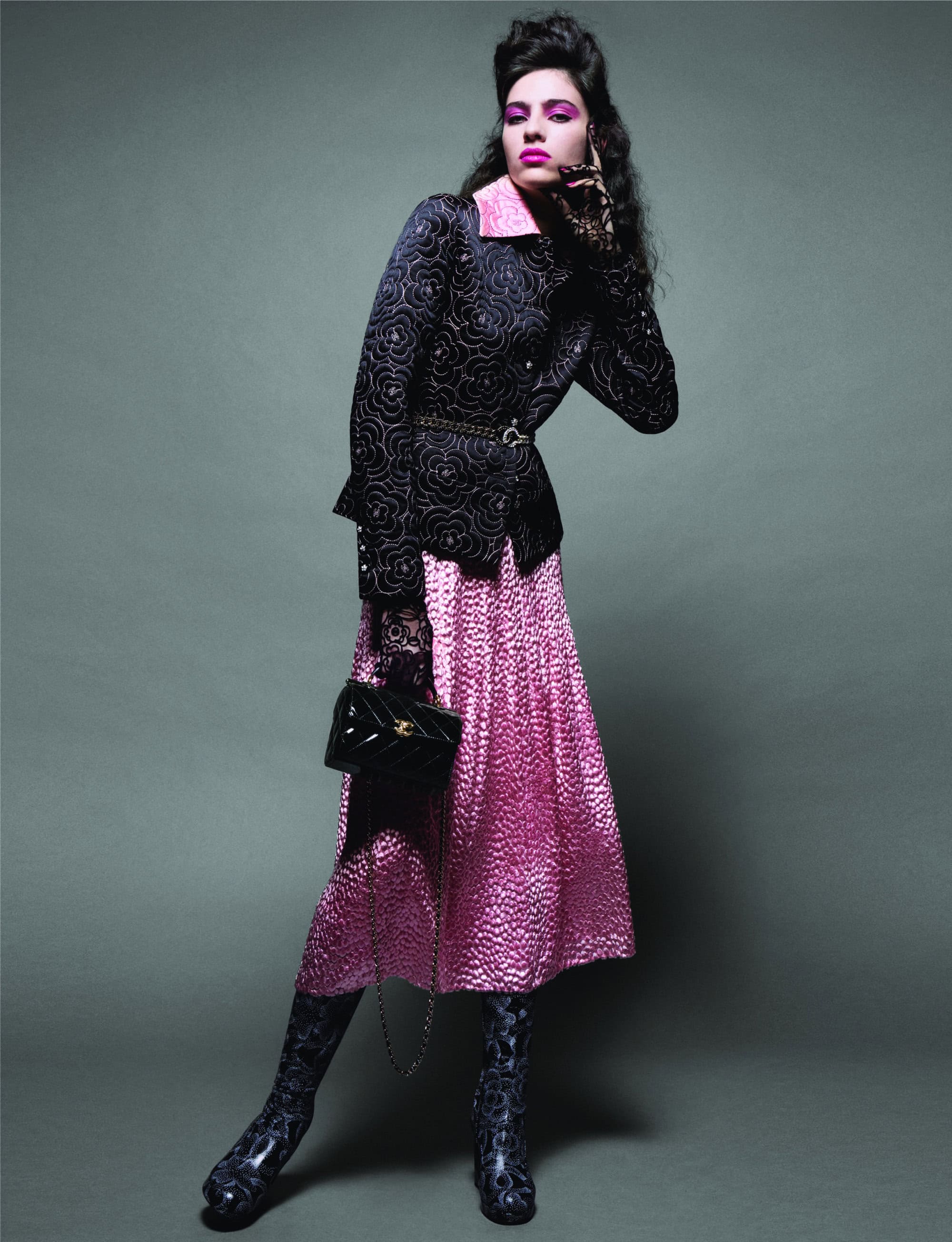 Fashionable woman in printed jacket and skirt, gloves, boots, and purse poses against a grey backdrop in a studio shot.