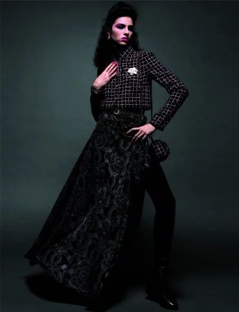 A woman in a black-and-white checkered jacket and long black skirt poses in a studio with low-key lighting and a dark-green background.