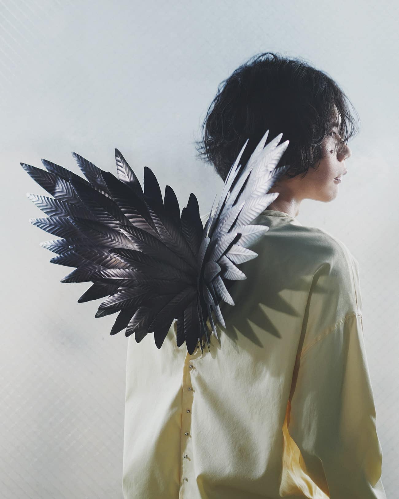 Profile shot of a person with short dark hair wearing an off-white shirt with large, dark feathered wings attached to the back.