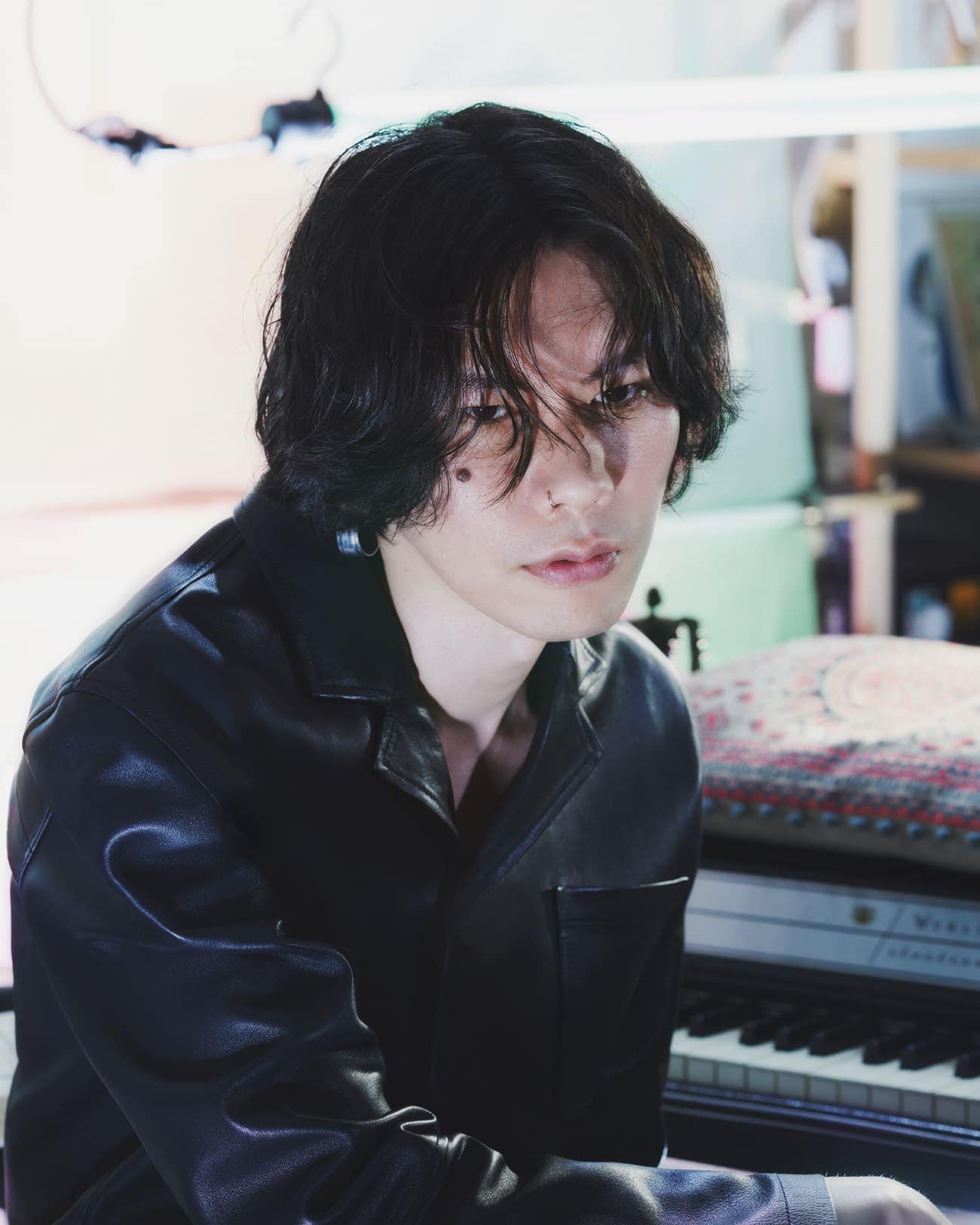 Portrait of a young adult with dark hair wearing a black jacket indoors, seated at a piano with soft, diffused lighting.