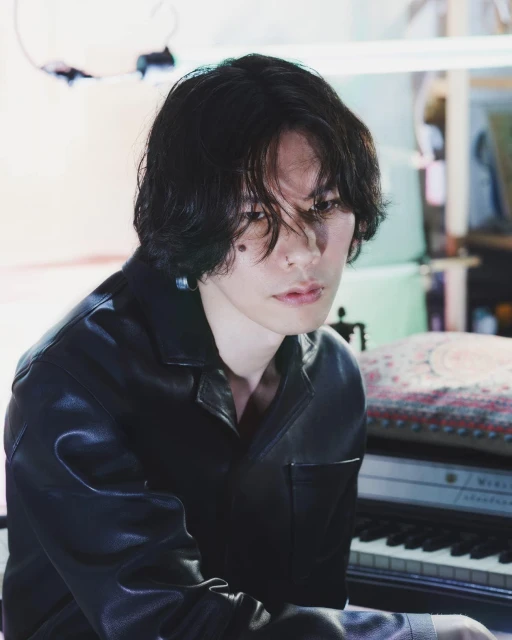 Portrait of a young adult with dark hair wearing a black jacket indoors, seated at a piano with soft, diffused lighting.