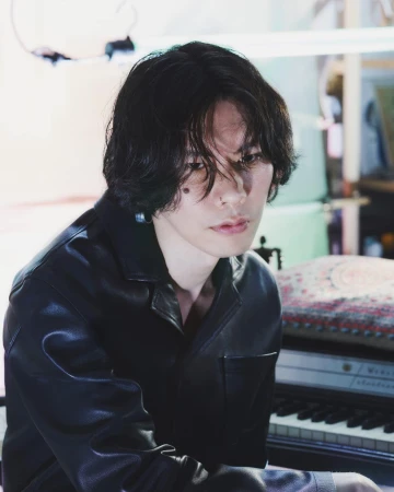 Portrait of a young adult with dark hair wearing a black jacket indoors, seated at a piano with soft, diffused lighting.