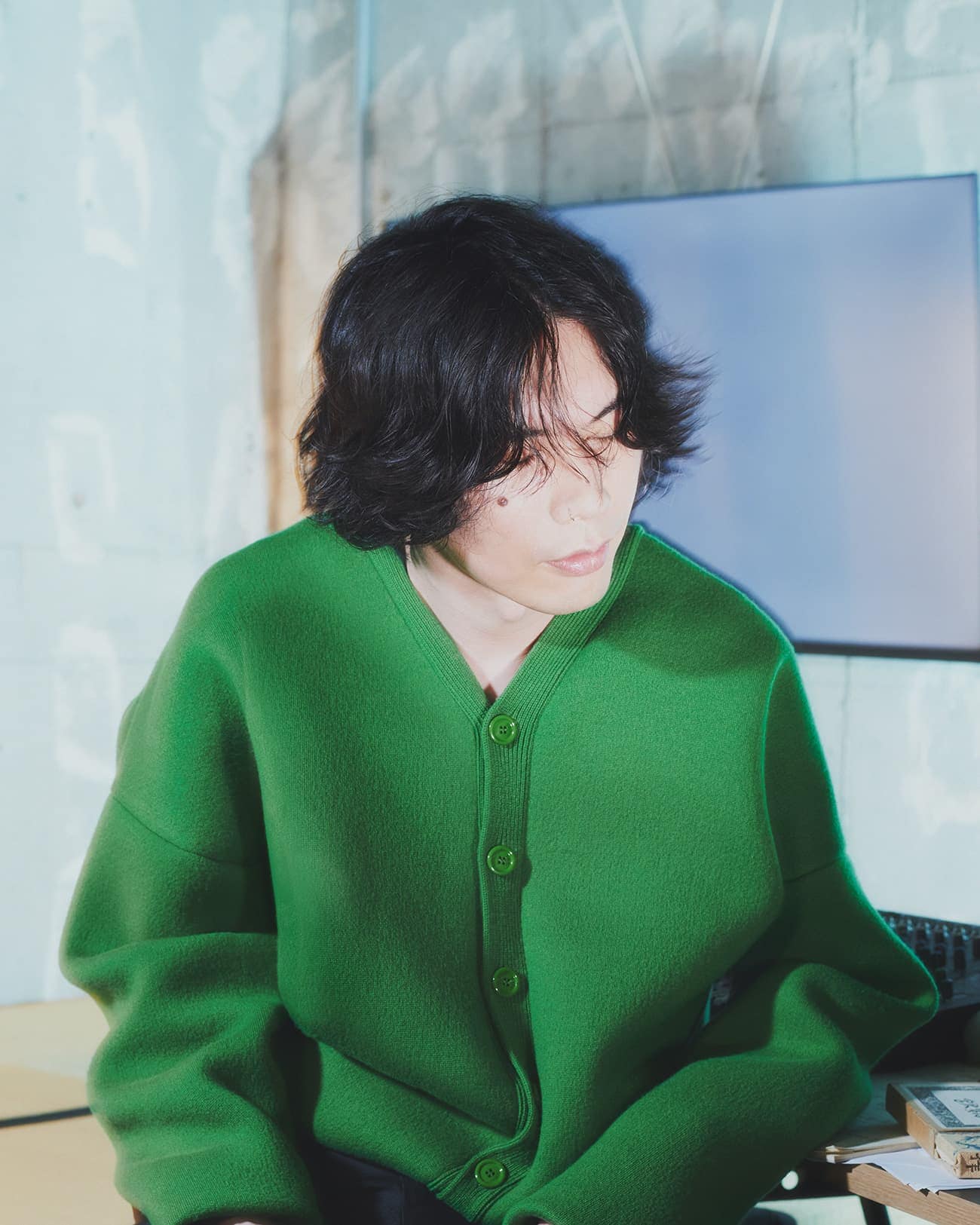 An adult with long dark hair and fair skin is wearing a green cardigan indoors, sitting in front of a monitor in a minimalist space.