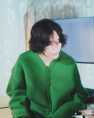 An adult with long dark hair and fair skin is wearing a green cardigan indoors, sitting in front of a monitor in a minimalist space.