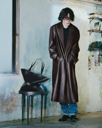 Kenshi Yonezu poses in a long dark brown leather coat beside a black Loewe geometric tote bag resting on a stool.