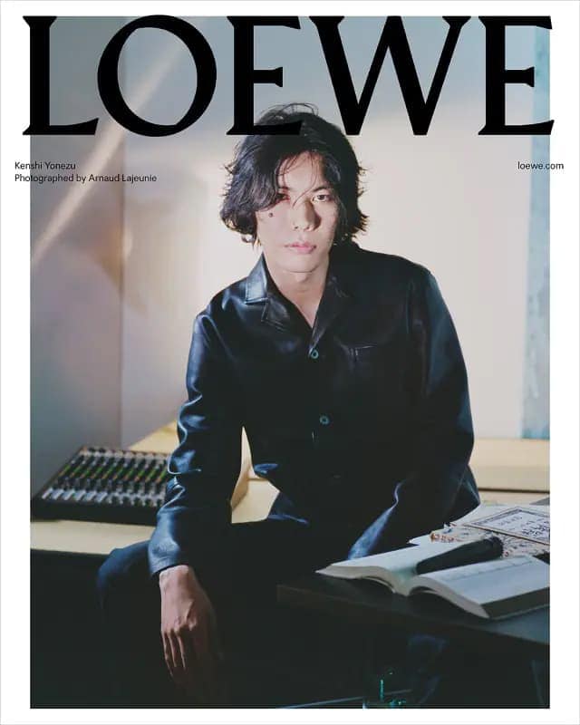 An Asian man with long, dark hair wearing a dark leather shirt posing indoors next to a table with a sound mixer and a book.