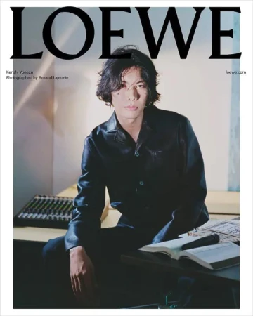 An Asian man with long, dark hair wearing a dark leather shirt posing indoors next to a table with a sound mixer and a book.