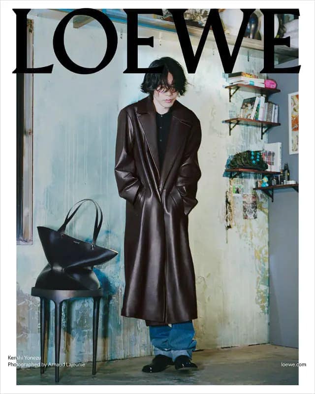 Editorial fashion shot: Young man with dark hair in brown leather coat stands indoors with a black tote bag on a black side table.