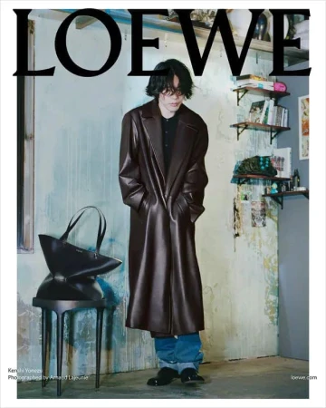 Editorial fashion shot: Young man with dark hair in brown leather coat stands indoors with a black tote bag on a black side table.