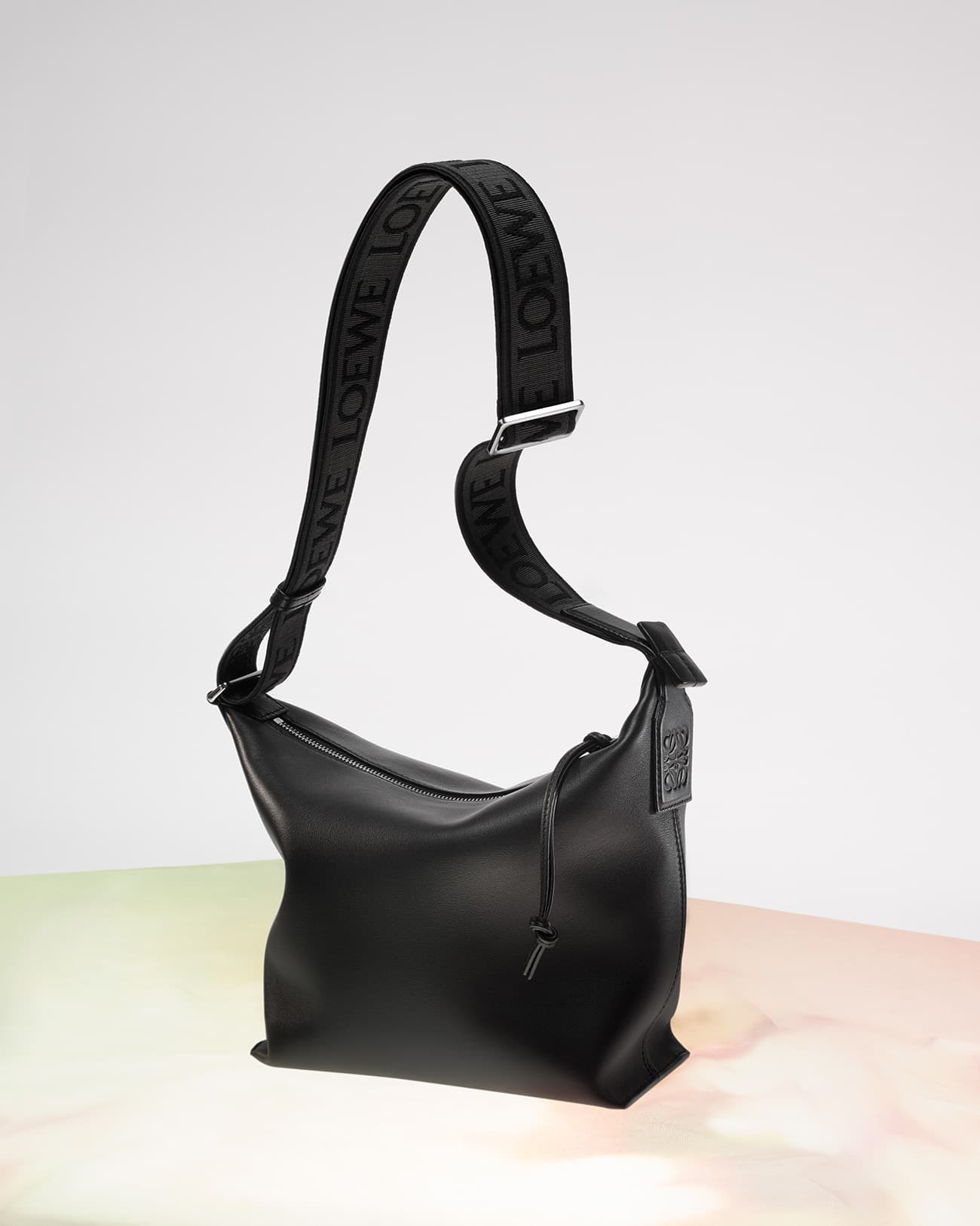 Studio shot of a black leather Loewe handbag with a patterned shoulder strap on a gradient background, showcasing its sleek design.