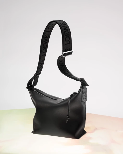 Studio shot of a black leather Loewe handbag with a patterned shoulder strap on a gradient background, showcasing its sleek design.