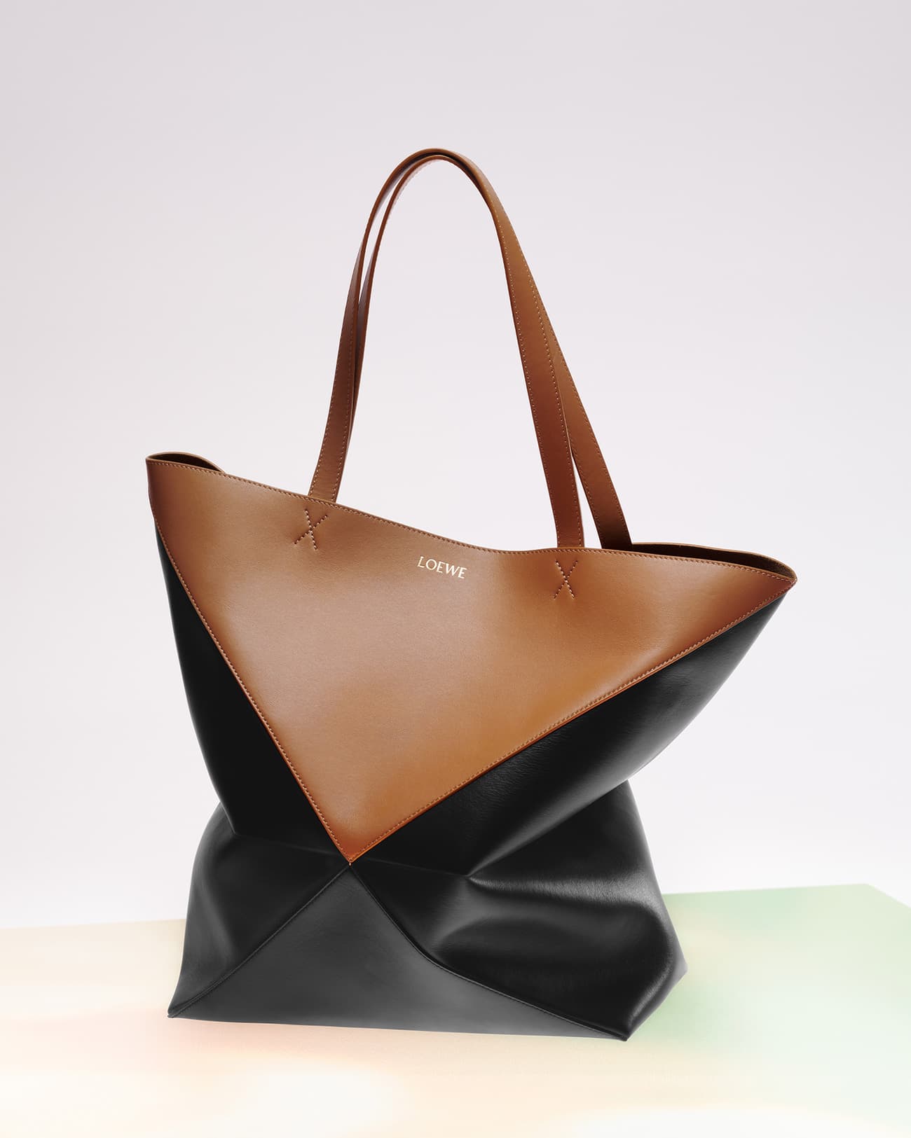Tan and black leather Loewe tote bag with top handles, photographed on a gradient white background in a studio setting.