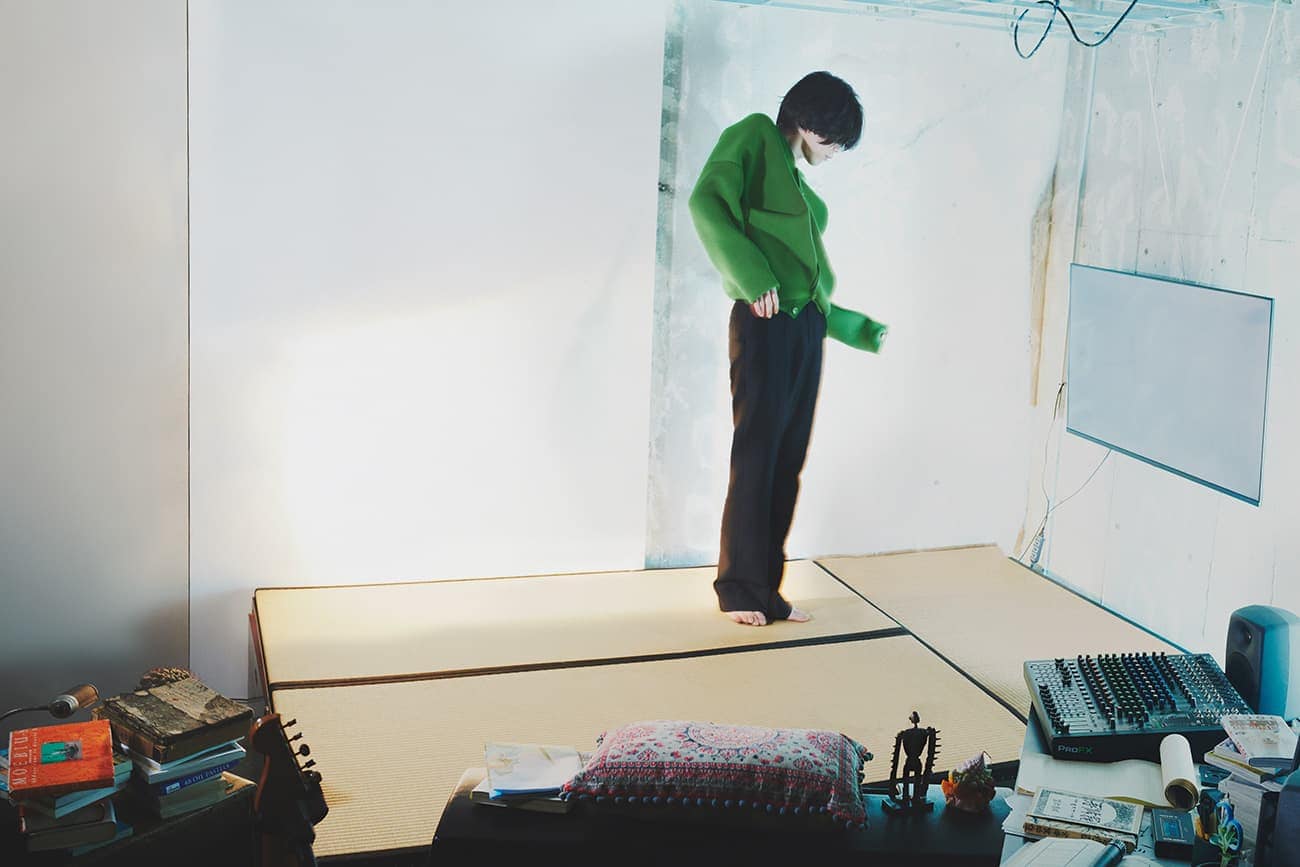 A barefoot adult stands in a casual green top and black pants in a room with light walls and a desk with books and music equipment.
