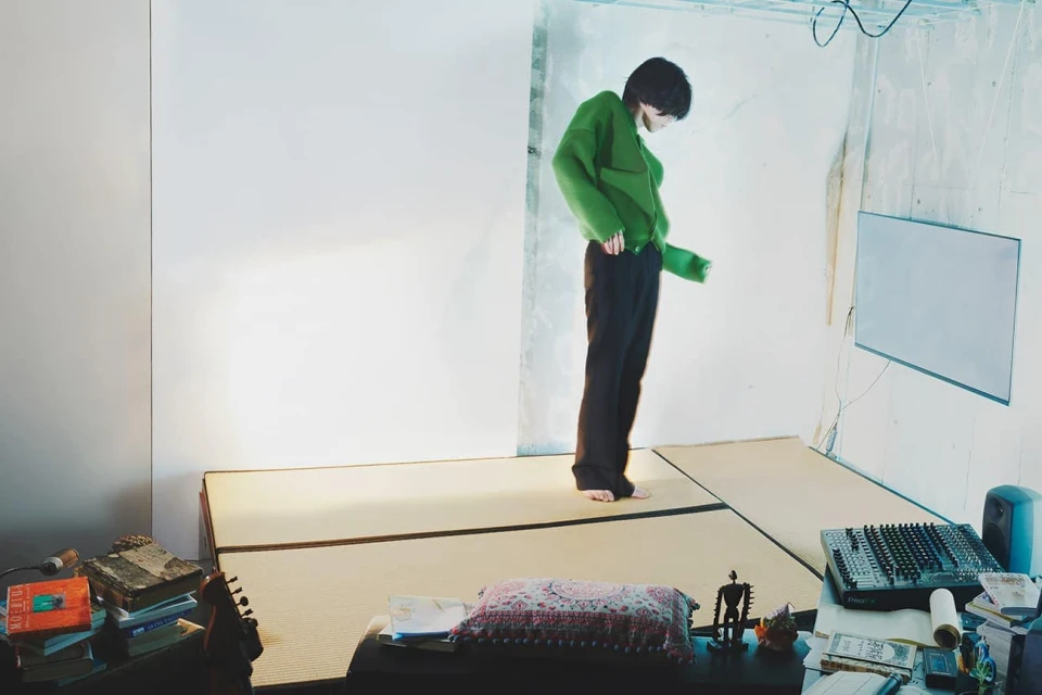 A barefoot adult stands in a casual green top and black pants in a room with light walls and a desk with books and music equipment.