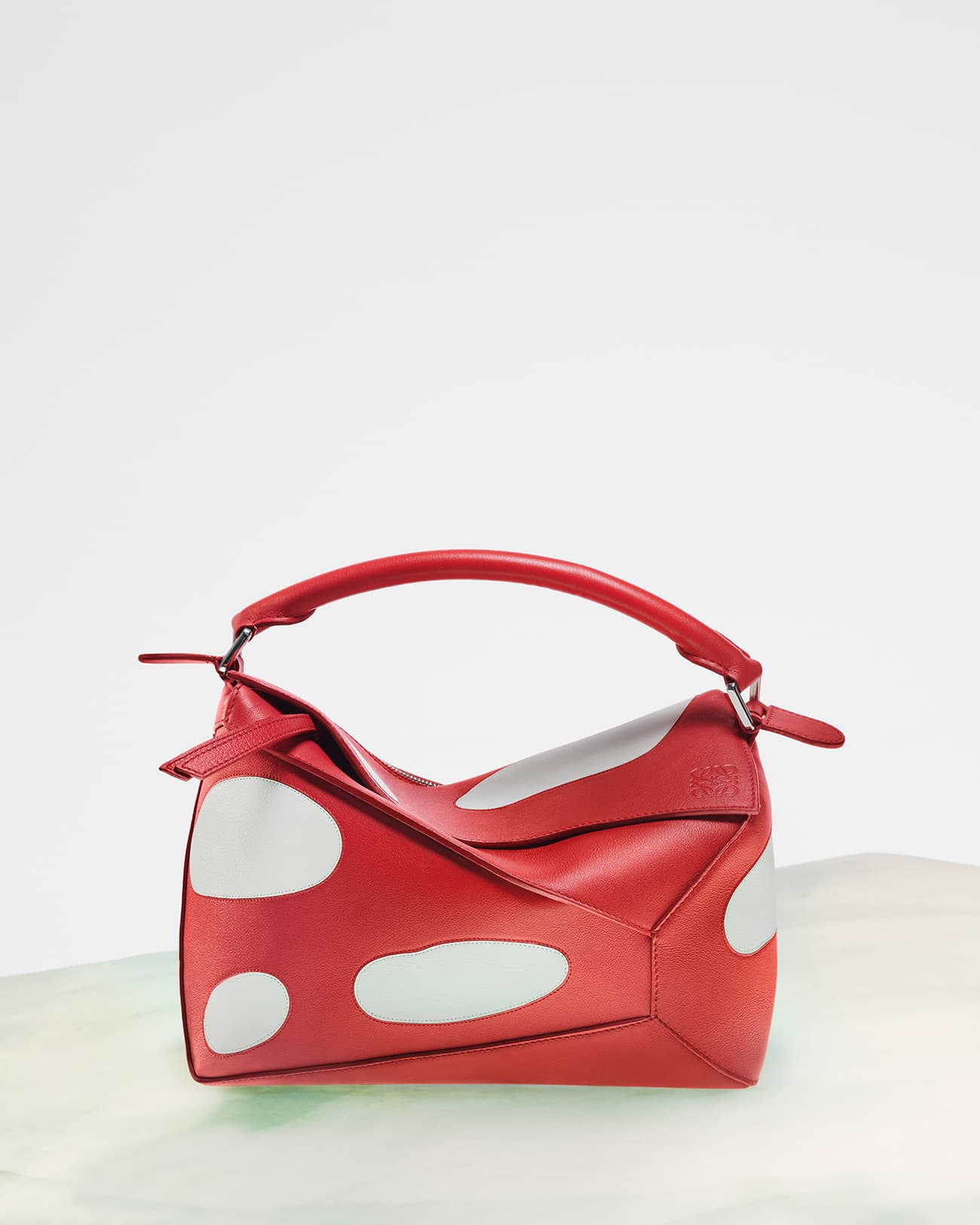 Elevated product shot of a red leather Loewe Puzzle bag with white polka dots on a simple green and white surface.