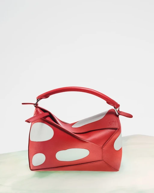 Elevated product shot of a red leather Loewe Puzzle bag with white polka dots on a simple green and white surface.