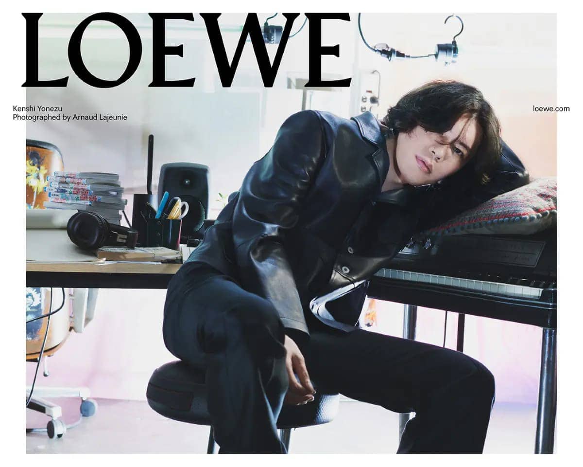 Adult male fashion model poses in a leather jacket leaning on a piano next to a desk with audio and video equipment in a studio.