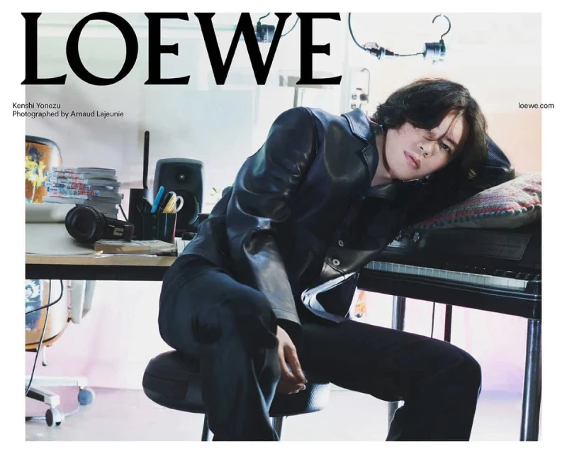 Adult male fashion model poses in a leather jacket leaning on a piano next to a desk with audio and video equipment in a studio.