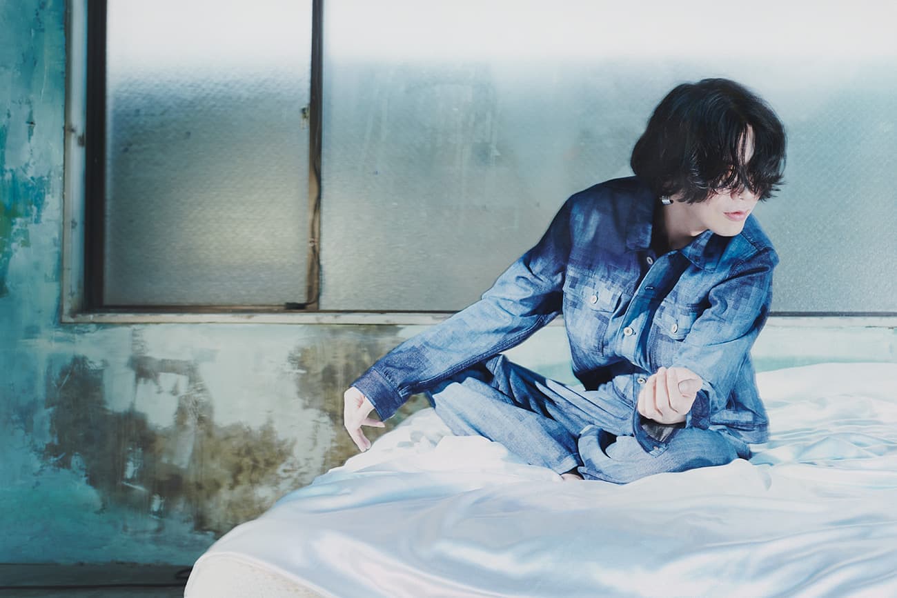 Young adult in a blue denim outfit sits cross-legged on a bed in a sparse room with a window, posing moodily, capturing introspective portrait.