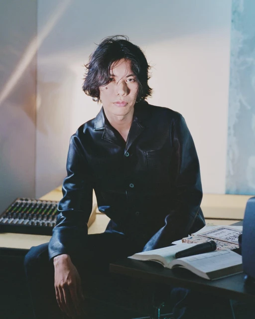 Portrait of a young Asian man in a black leather jacket sitting at a table with music equipment in a softly lit room.