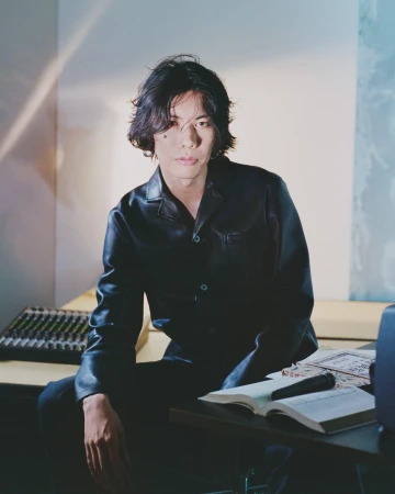 Portrait of a young Asian man in a black leather jacket sitting at a table with music equipment in a softly lit room.