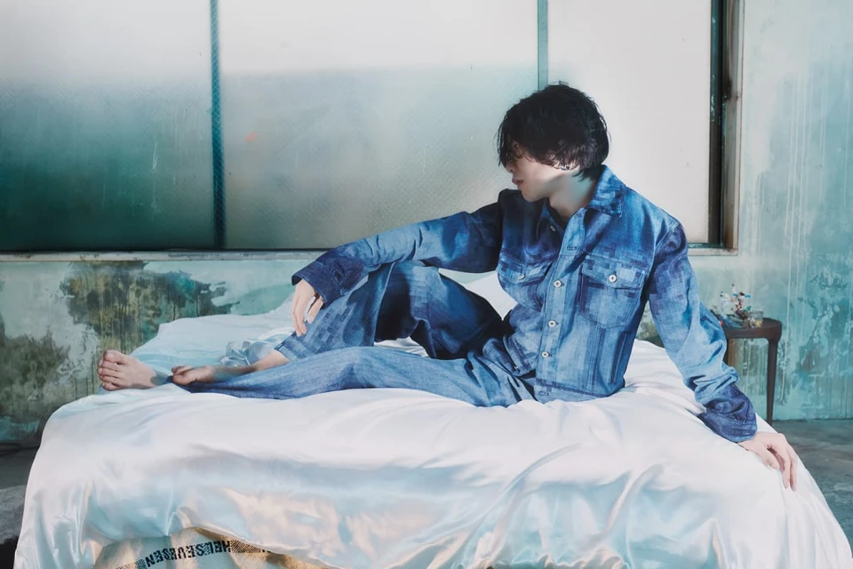 Adult male sits on a bed in denim clothing, looking off to the side, with a glass window and distressed wall in the background.