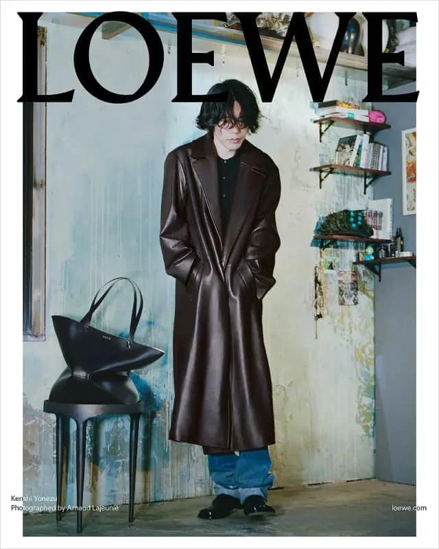 Kenshi Yonezu wearing a long brown leather coat and blue jeans standing in front of a textured wall for Loewe Fall 2023 campaign.