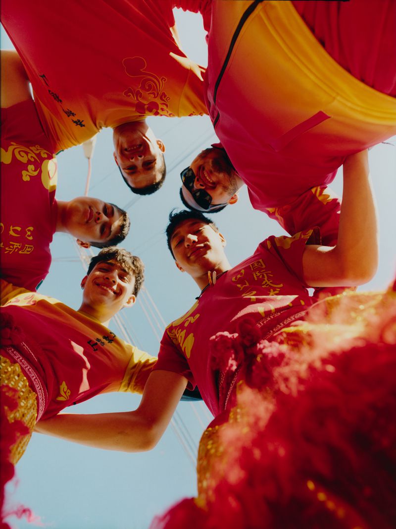 Group of five smiling men in red and yellow shirts looking down at the camera in a circle, low angle shot, clear blue sky.
