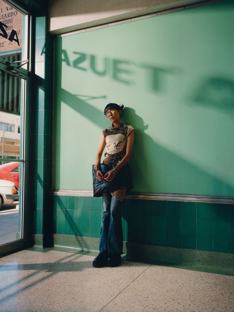 Young woman with glasses leans against a green tiled wall with a shadow. She wears jeans and a patterned tank top.