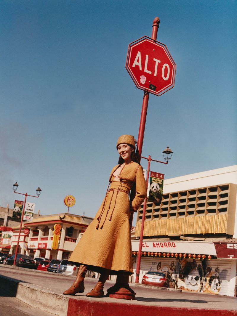 Woman in tan coat poses next to an Alto street sign in an outdoor setting. The building and cars are visible in the background.