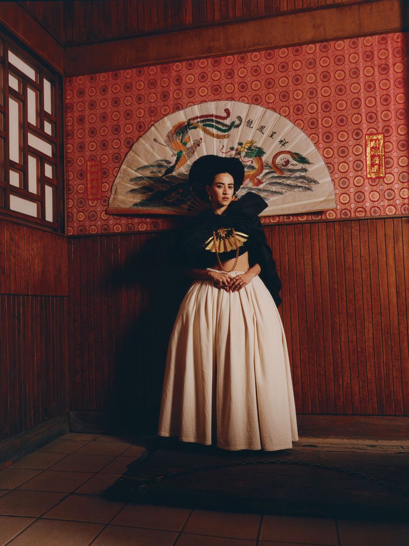 Portrait of an adult female in a traditional costume, featuring a large skirt, black cape, and decorative fan, set against a patterned wall.