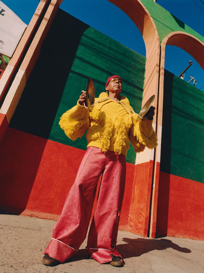 Adult male stands outdoors in a yellow sweater, pink pants, and a red headscarf while holding cymbals in front of a colorful wall.