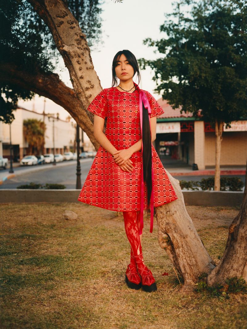 A young woman in a red dress stands in a park near a tree, showcasing an editorial fashion style with a blurred urban background.