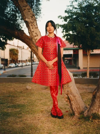 A young woman in a red dress stands in a park near a tree, showcasing an editorial fashion style with a blurred urban background.