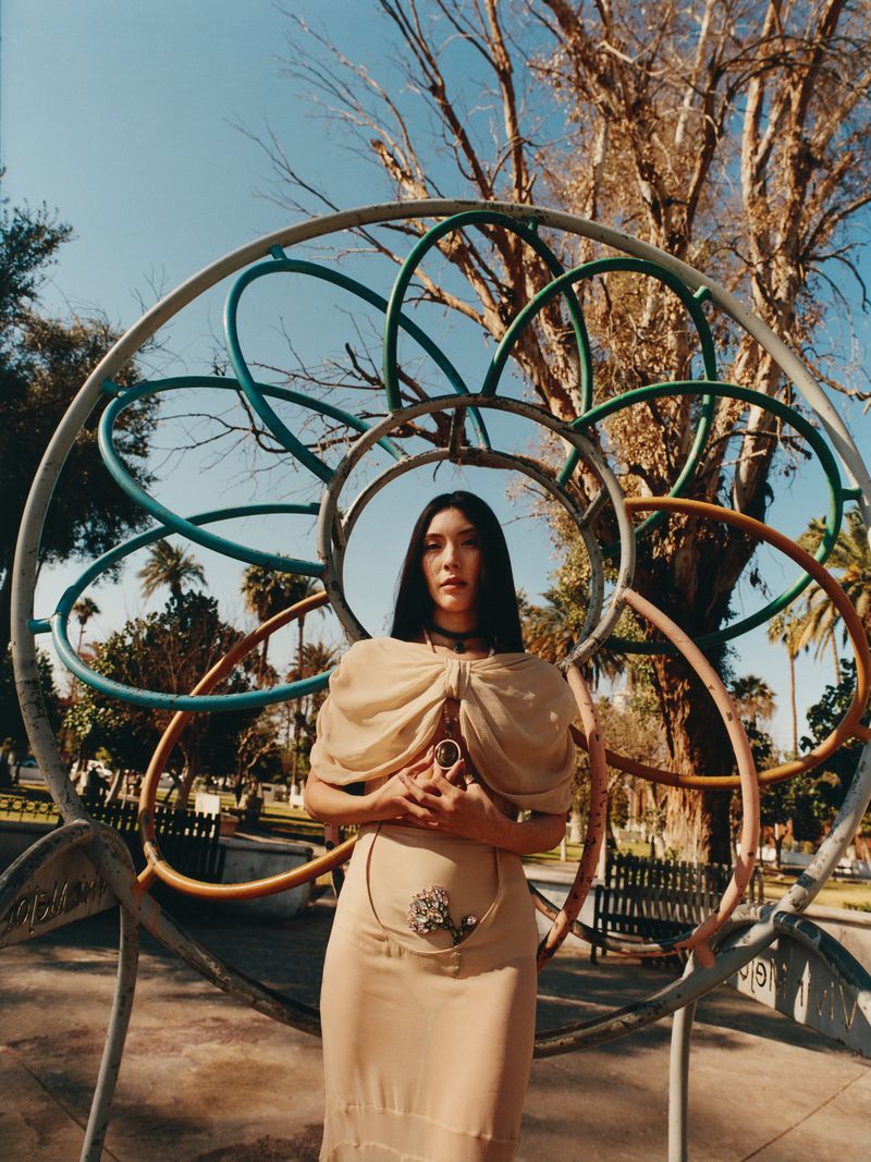 Woman in beige dress poses in front of colorful metal playground structure in sunny outdoor setting with trees and clear blue sky.
