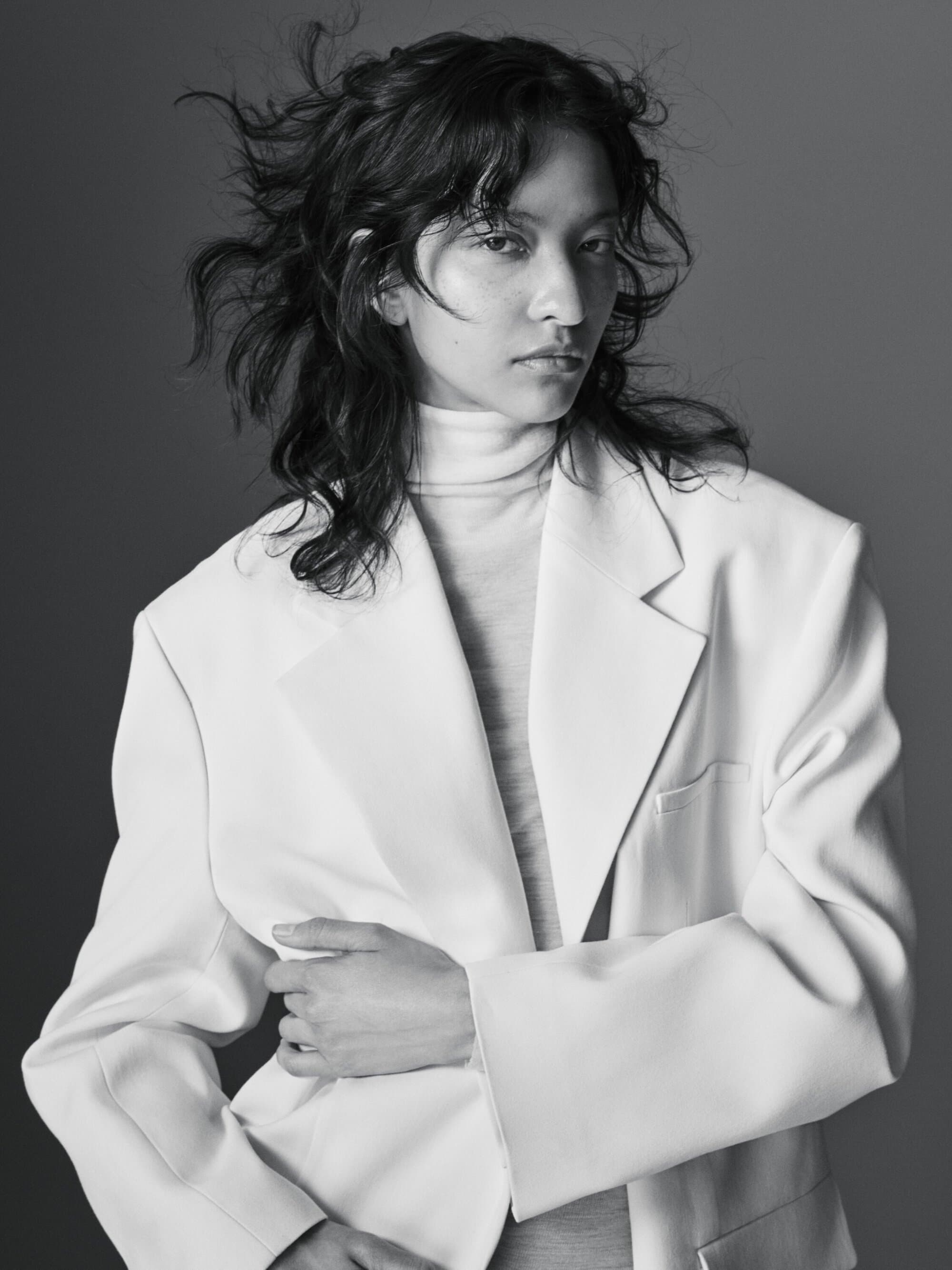 Portrait of a model with dark tousled hair wearing an oversized white blazer and turtleneck sweater against a grey backdrop