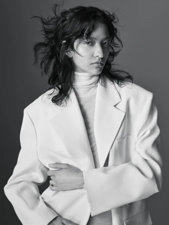Portrait of a model with dark tousled hair wearing an oversized white blazer and turtleneck sweater against a grey backdrop