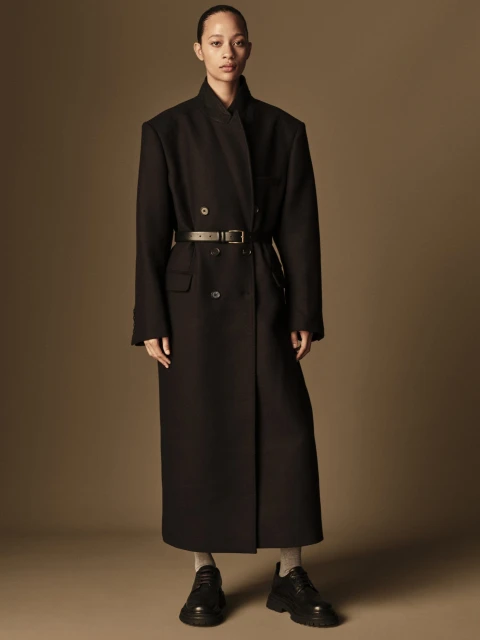 An adult female model poses in a long, dark belted coat and platform shoes in a full shot against a plain brown backdrop.