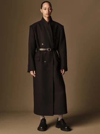 An adult female model poses in a long, dark belted coat and platform shoes in a full shot against a plain brown backdrop.