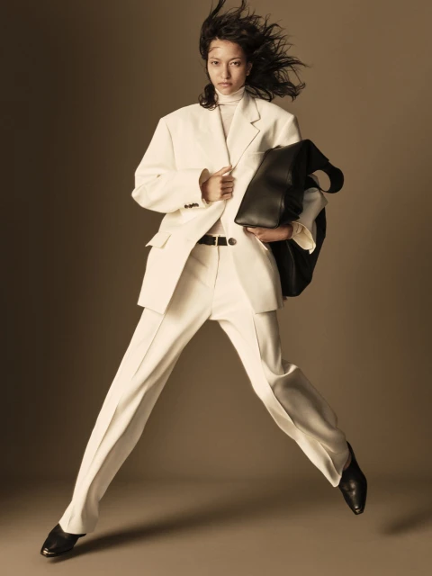 Asian woman in motion, wearing an oversized white suit and black leather tote bag against a neutral backdrop in a fashion setting.