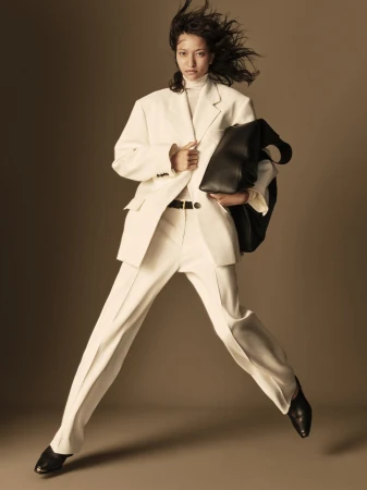 Asian woman in motion, wearing an oversized white suit and black leather tote bag against a neutral backdrop in a fashion setting.