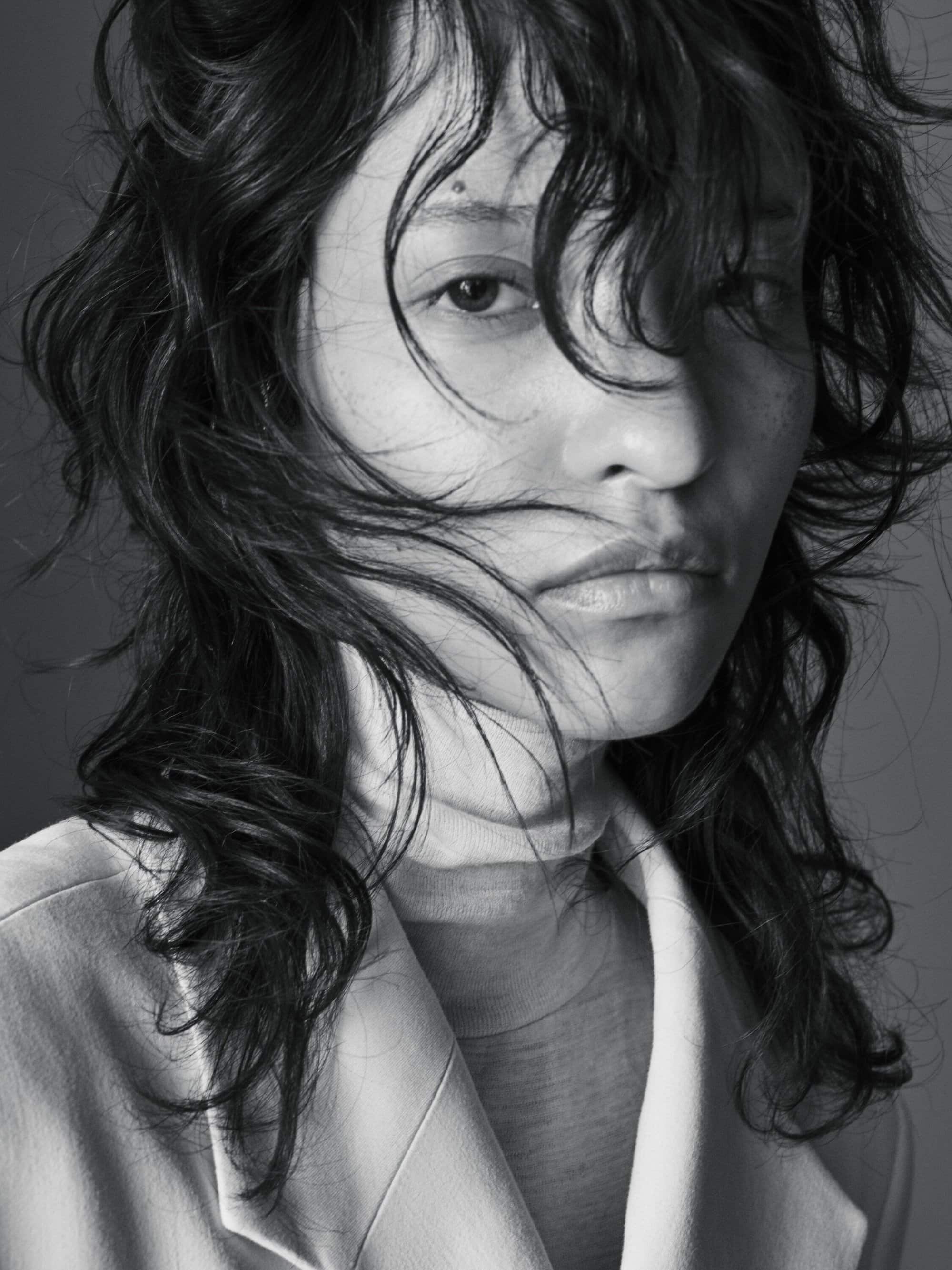 Close-up portrait of a woman with tousled dark hair wearing a beige blazer against a plain gray studio background.