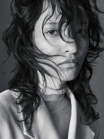 Close-up portrait of a woman with tousled dark hair wearing a beige blazer against a plain gray studio background.