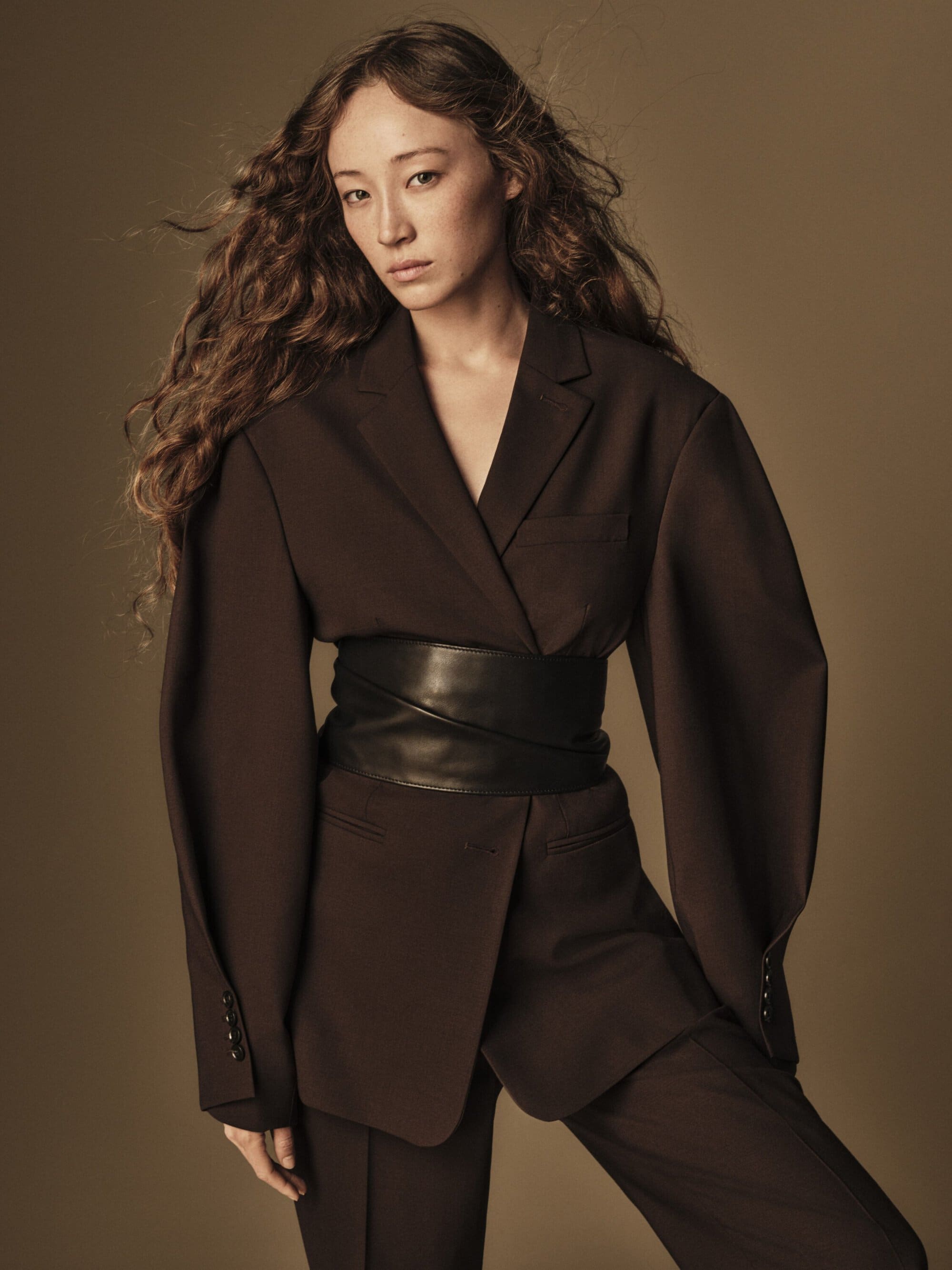 Portrait of an adult female with long, wavy brown hair, wearing a brown suit with a wide black belt against a neutral background.