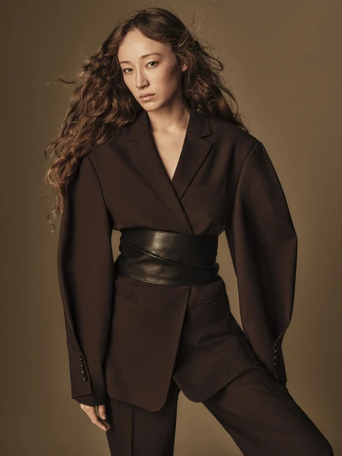 Portrait of an adult female with long, wavy brown hair, wearing a brown suit with a wide black belt against a neutral background.