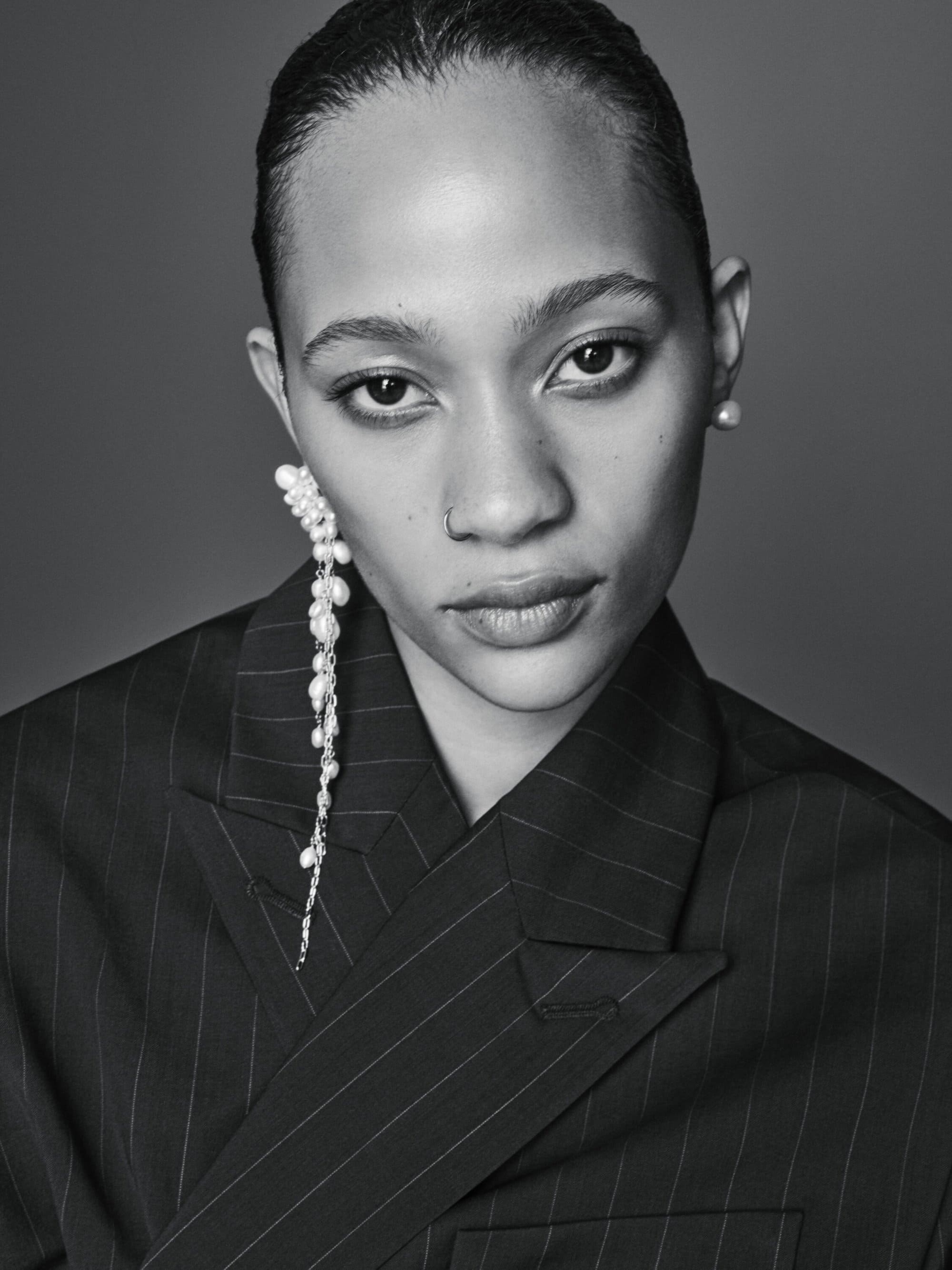 Striking black and white portrait of a woman with pearl earrings, wearing a pinstripe blazer against a dark grey backdrop.