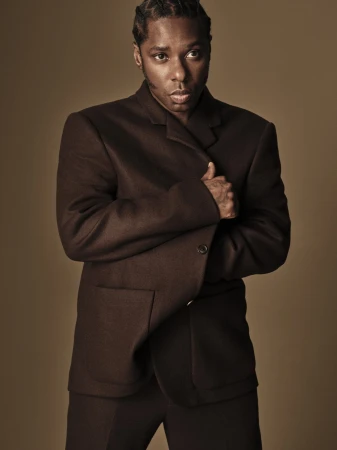 Curtis Harding posing in a dark brown tailored wool blazer against a warm neutral studio backdrop.