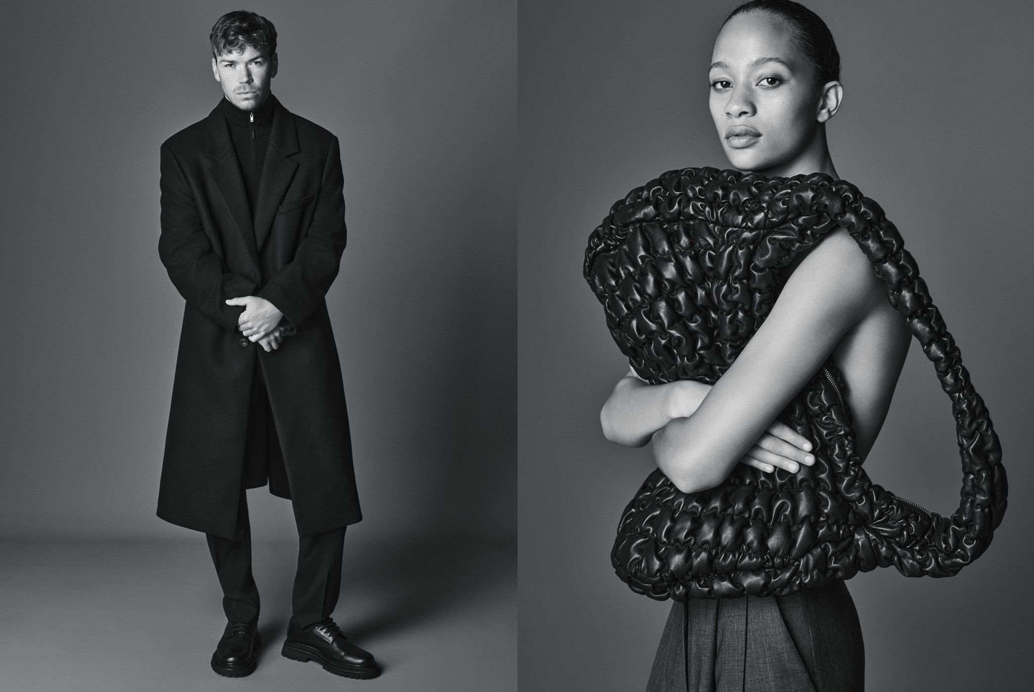 A man in a black wool coat and a woman holding a large black quilted bag against a neutral grey studio background in black and white.