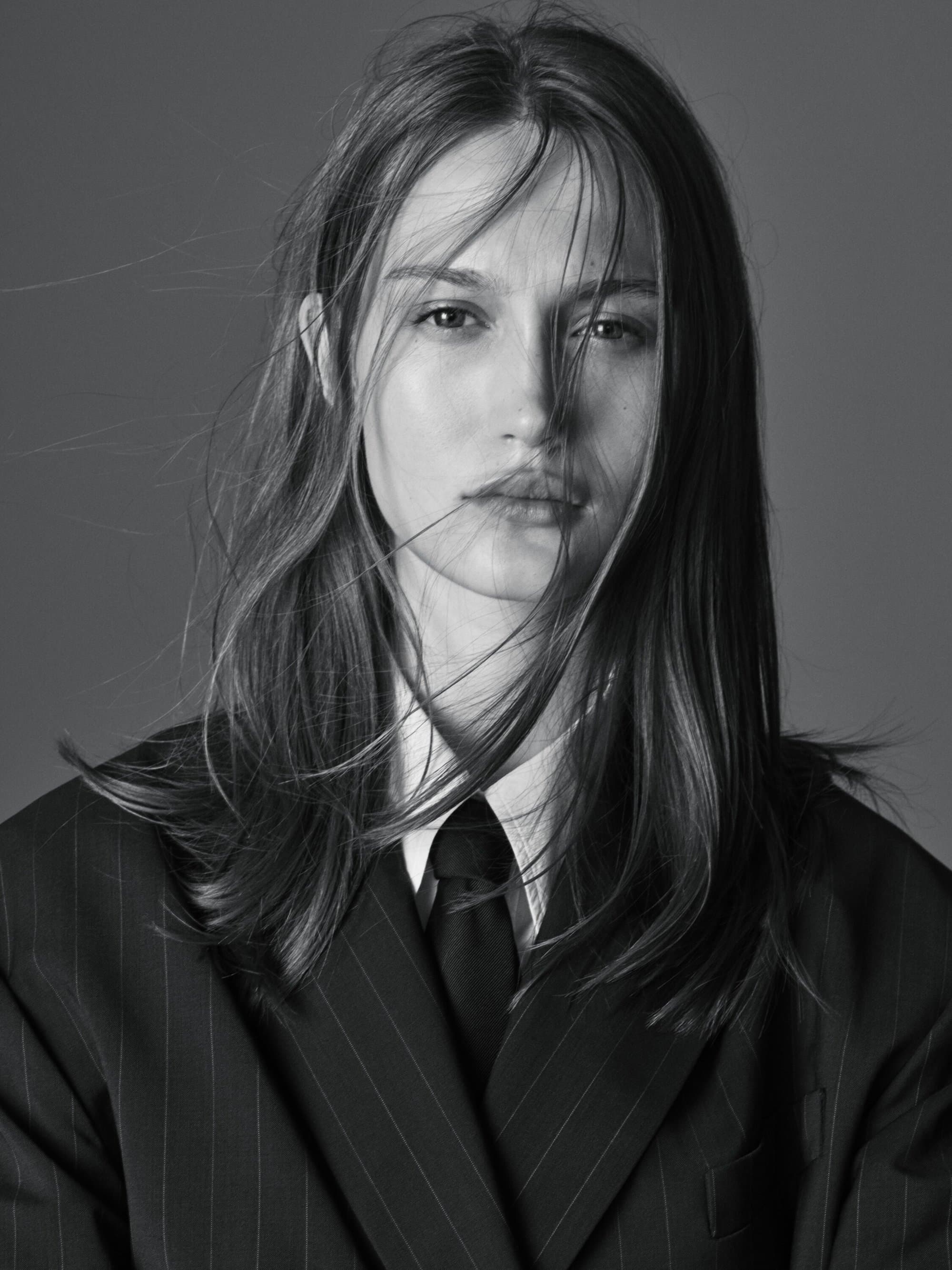 Monochrome portrait of a young woman with flowing hair, wearing a striped blazer, white collared shirt, and black tie, gazing at the camera.